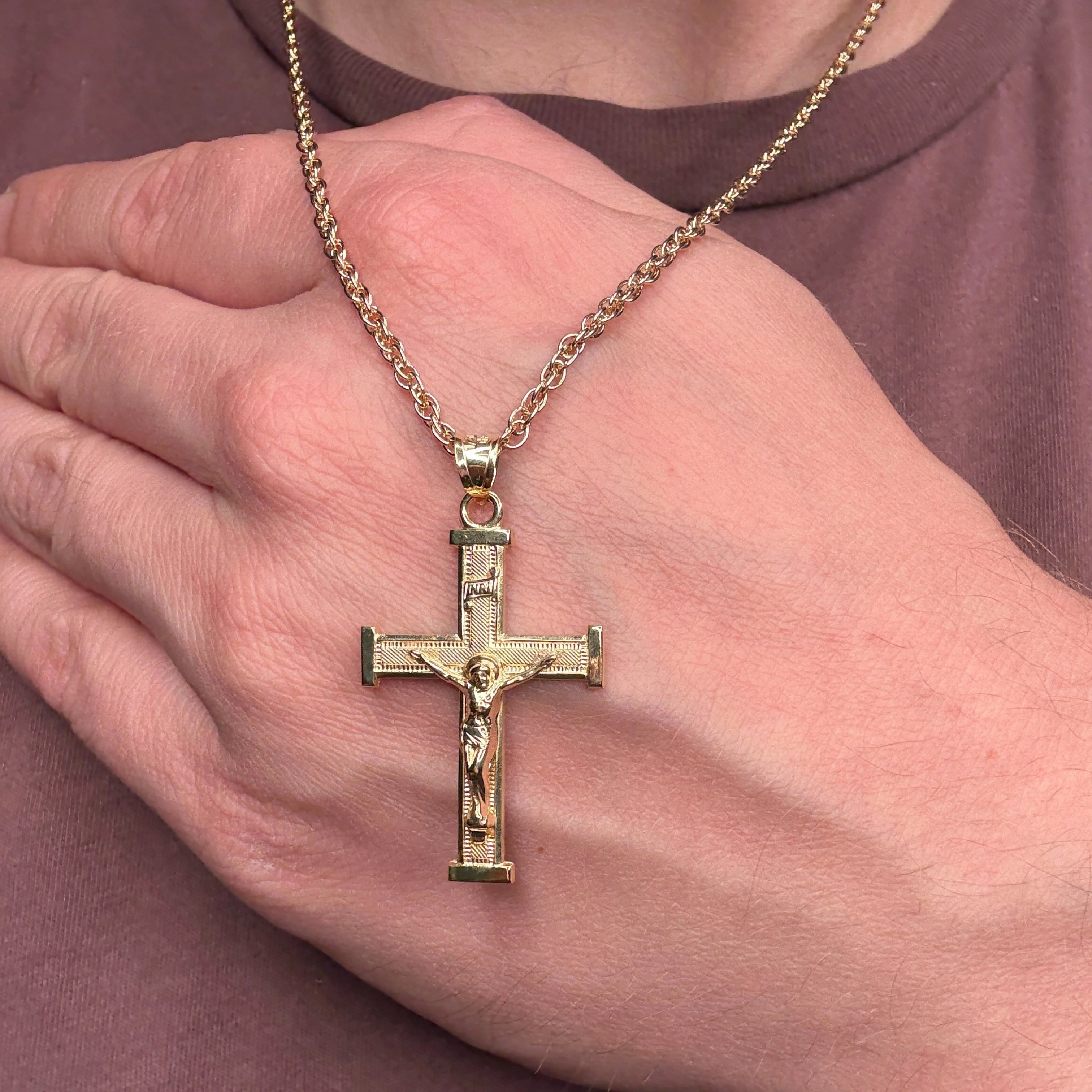 Gold crucifix pendant on a chain held in a hand against a plain background